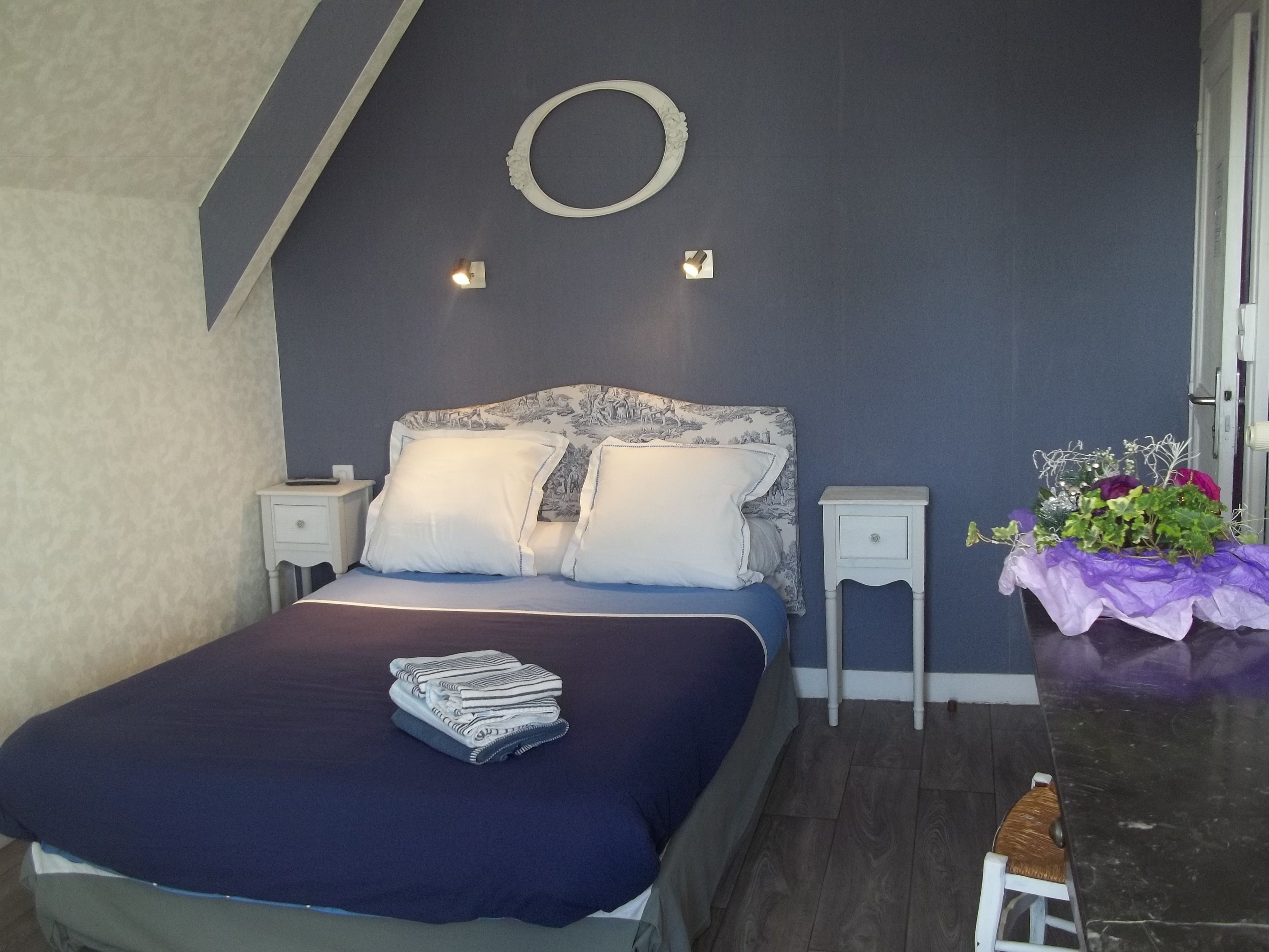 A bedroom with a bed, nightstands, a wall-mounted light fixture, a small table with a vase of flowers, and a wooden chair.