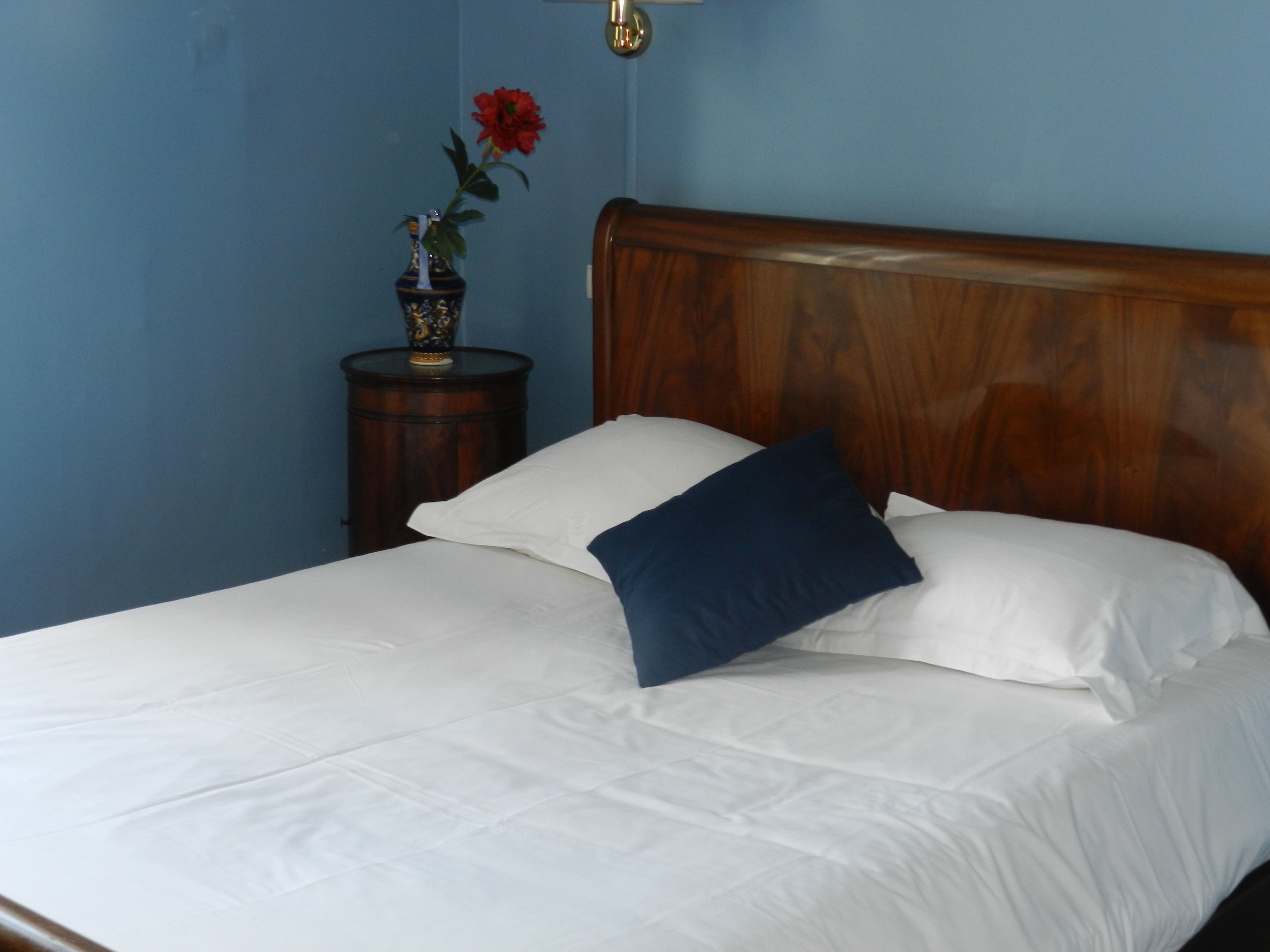 A neatly made bed with white linens and a blue pillow, a wooden headboard, and a small bedside table with a vase of red flowers.