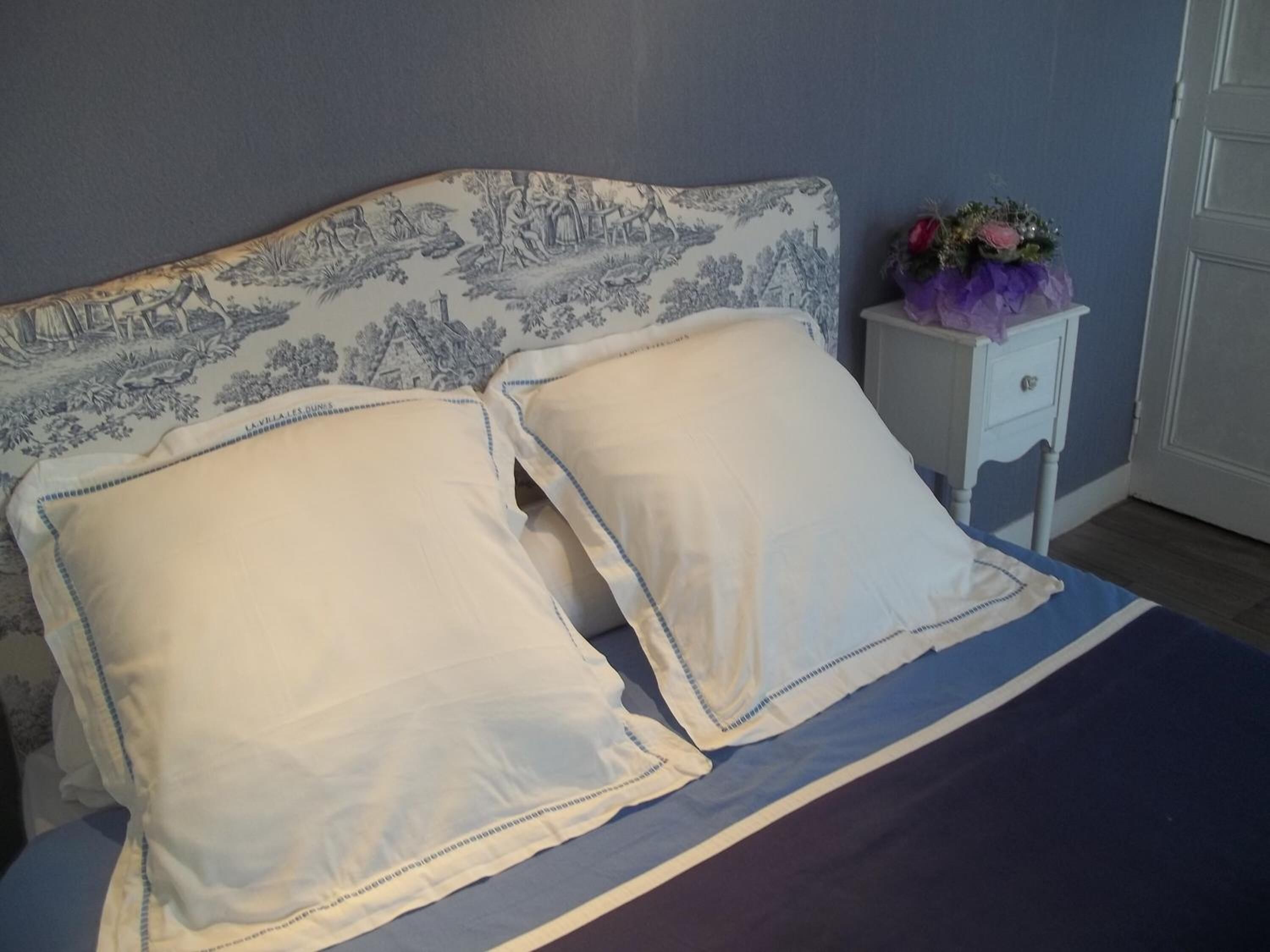 A bed with a patterned headboard, two pillows, a small bedside table with a vase of flowers, and a door in the background.