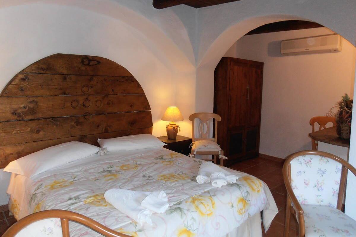 Quarto com cabeceira de madeira, cama com roupa de cama floral, mesa de cabeceira com abajur e duas cadeiras.
