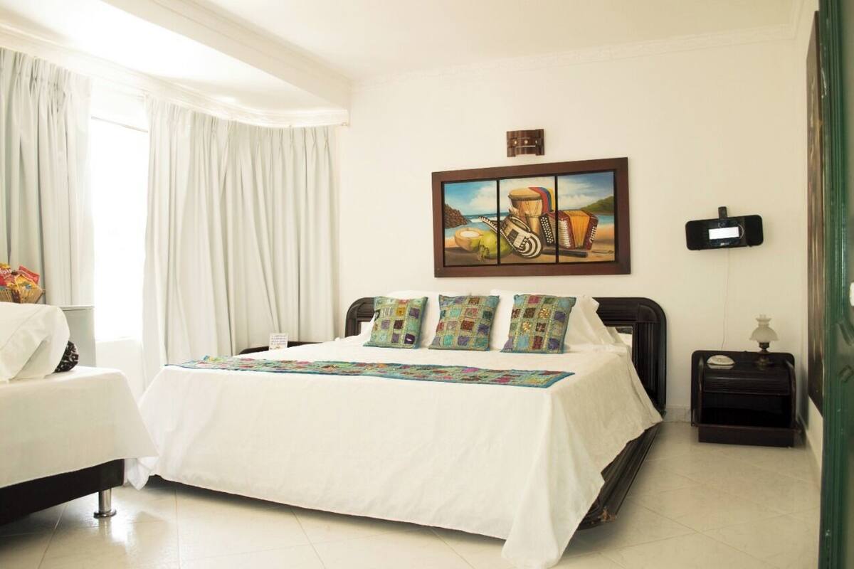 A hotel room with a large bed, a bedside table, a nightstand, a wall-mounted alarm clock, and a painting above the bed.