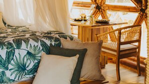 Una habitación con una cama, una mesa de comedor y una silla. La habitación tiene techo de paja y ventanas grandes.