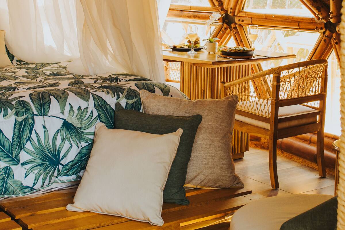A room with a bed, a dining table, and a chair. The room has a thatched roof and large windows.