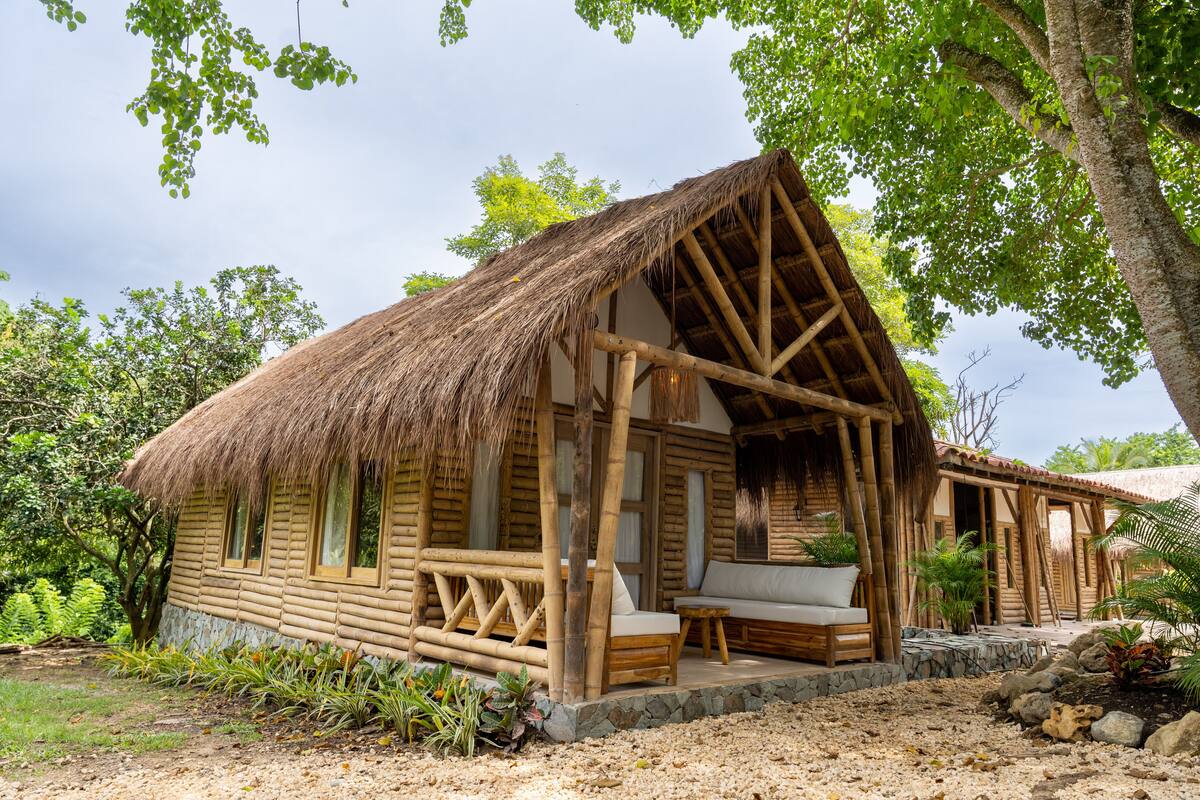 A wooden cabin with a thatched roof, a stone foundation, and a small porch with a bench.