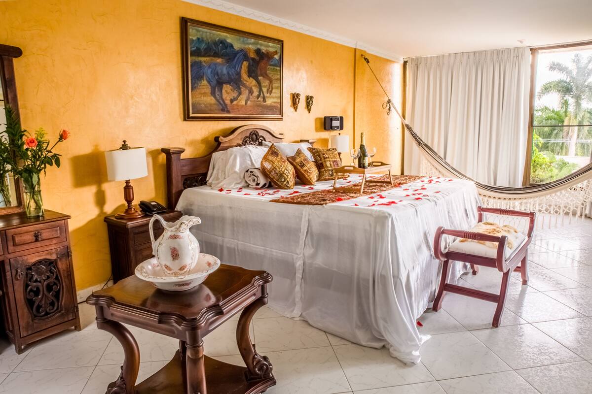A bedroom with a large bed, a hammock, a wooden nightstand, a table with a pitcher, and a chair.