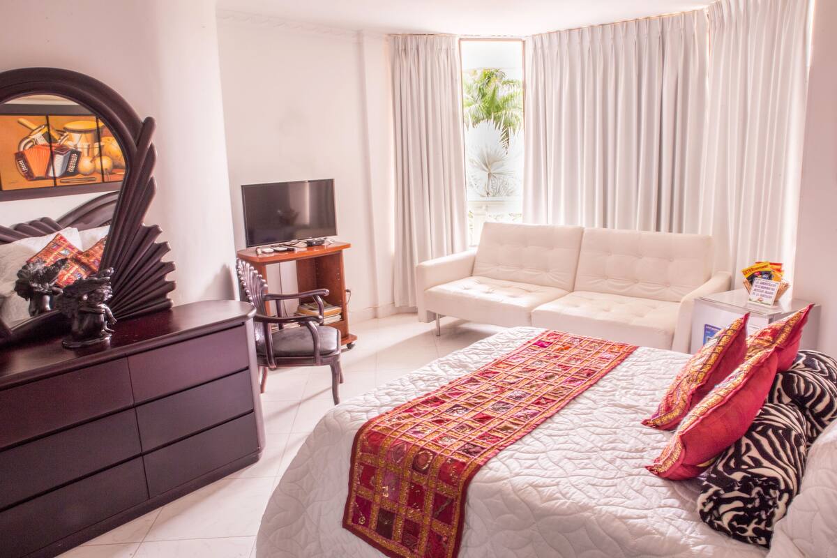 A hotel room with a bed, a sofa, a TV, a desk, and a window with curtains.