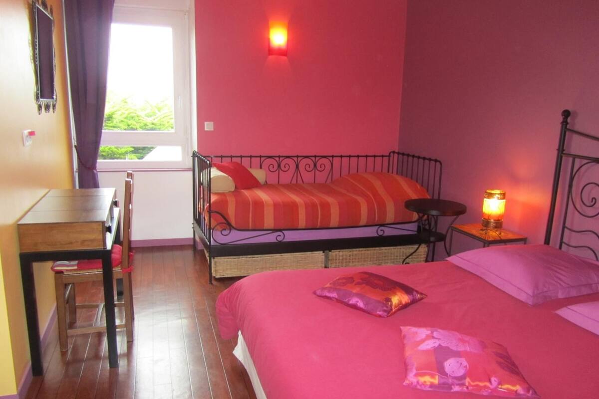 A bedroom with a bed, a desk, a chair, a nightstand with a lamp, and a window with curtains.