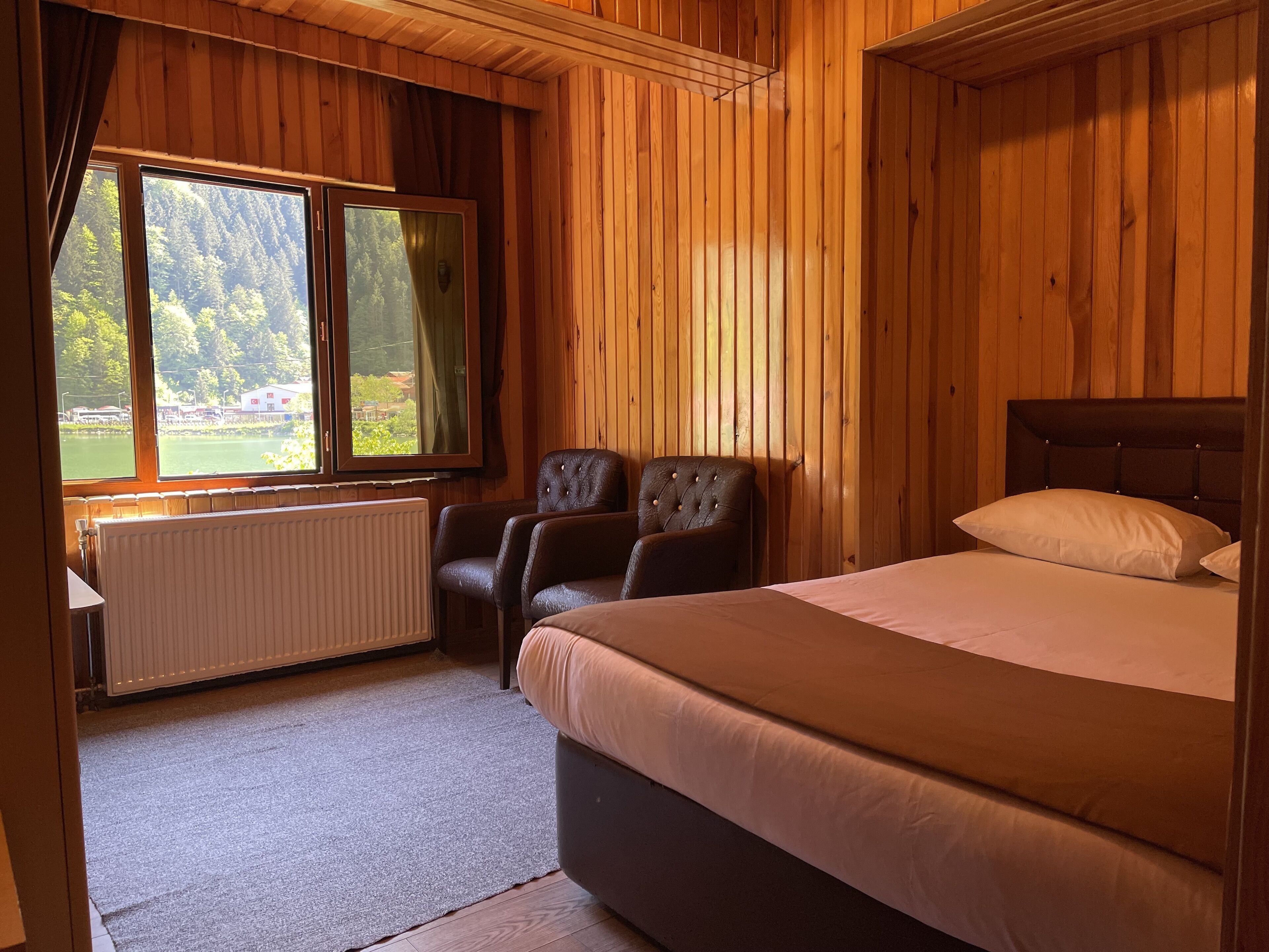 Habitación con paneles de madera, una cama, dos sillones, un radiador y ventana con vistas a zonas verdes y edificios.