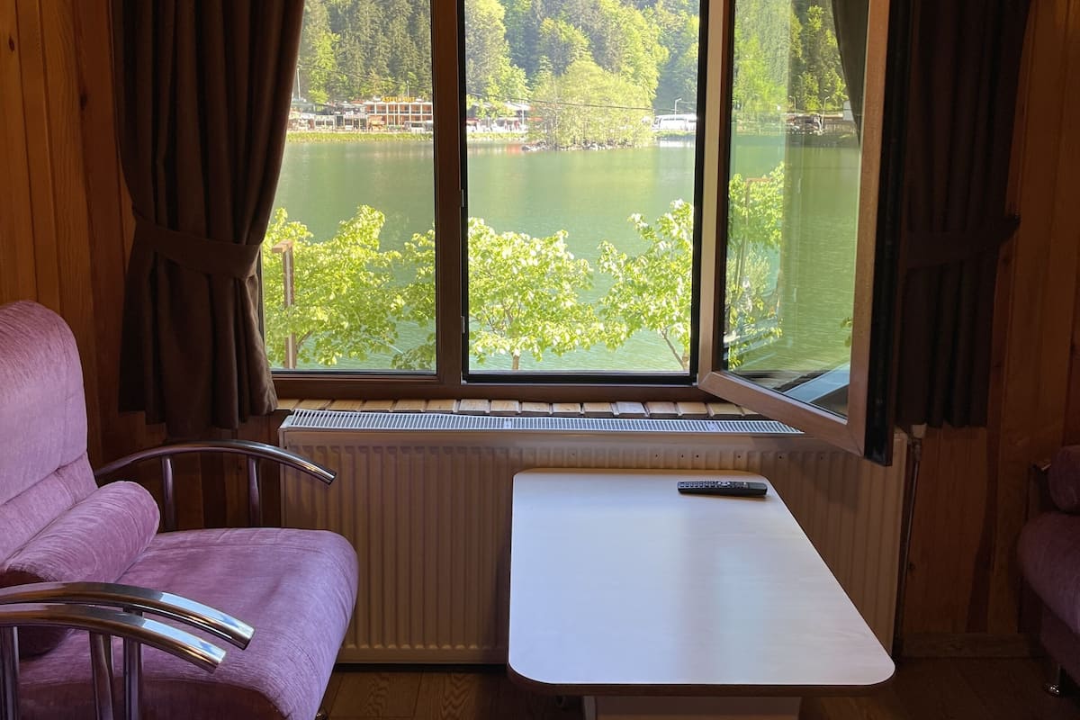 A cabin room with a wooden ceiling, a large window with curtains, a white table, and two purple chairs.