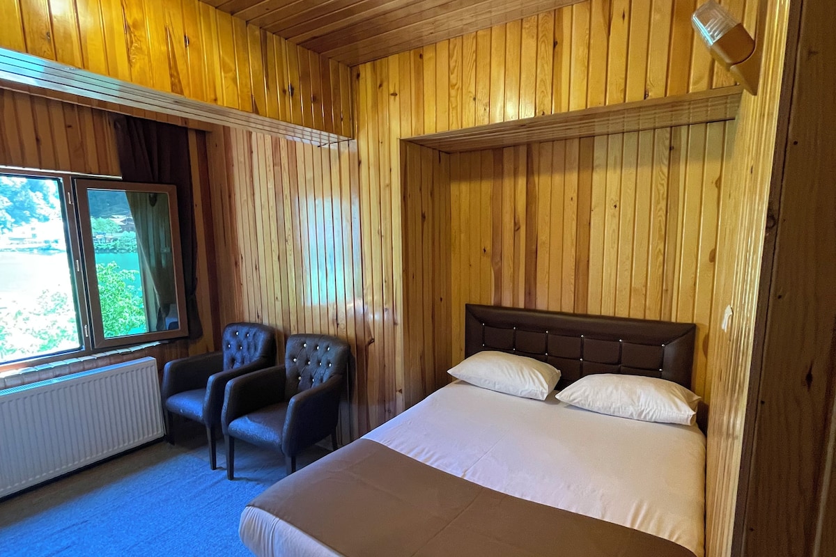 A wooden-paneled room with a bed, two armchairs, a radiator, and a window.
