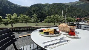 Una mesa con un plato de comida, una taza de té y una cesta de pan, con un lago y montañas al fondo.