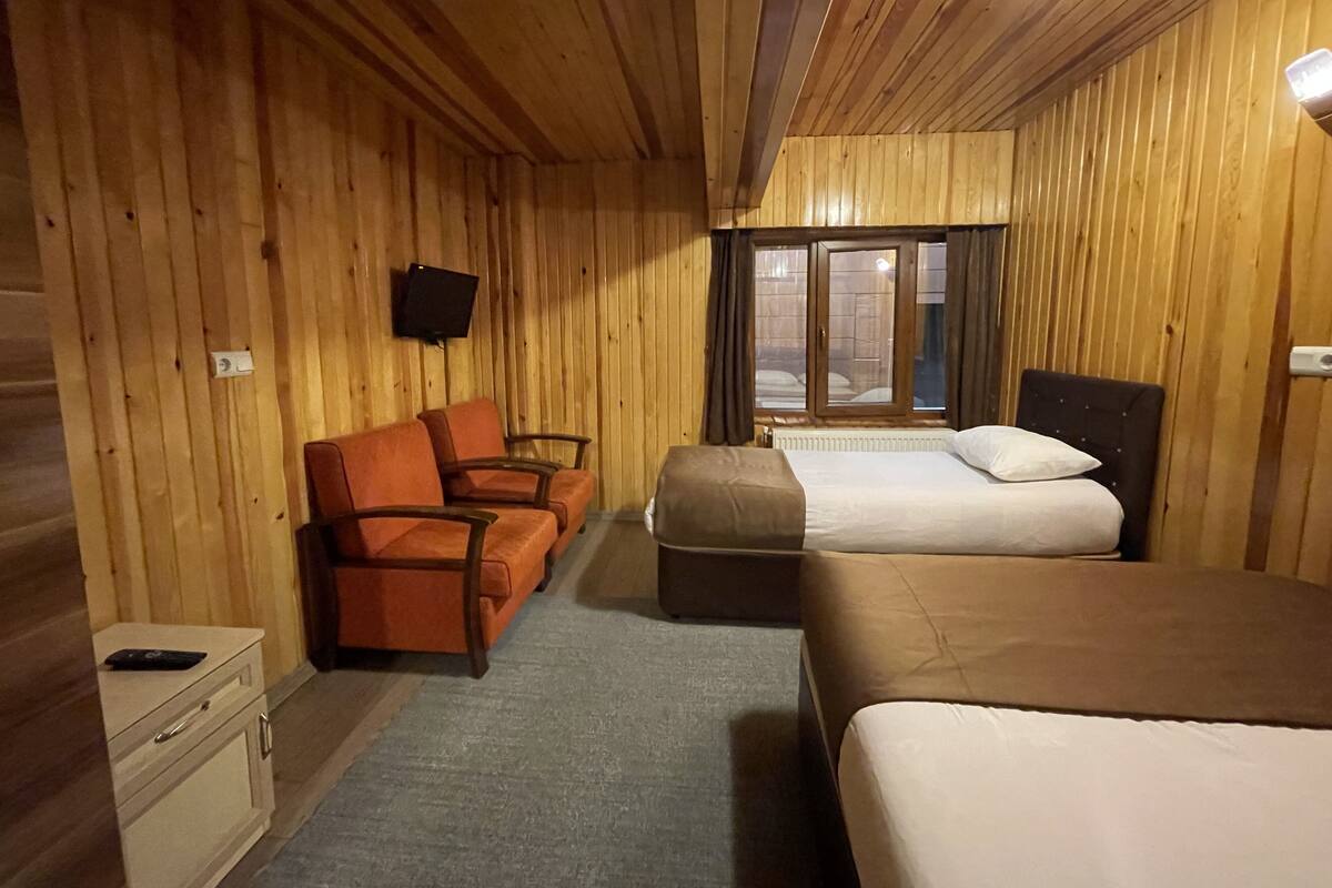 A wooden cabin room with two beds, a sofa, a TV, and a window with curtains.