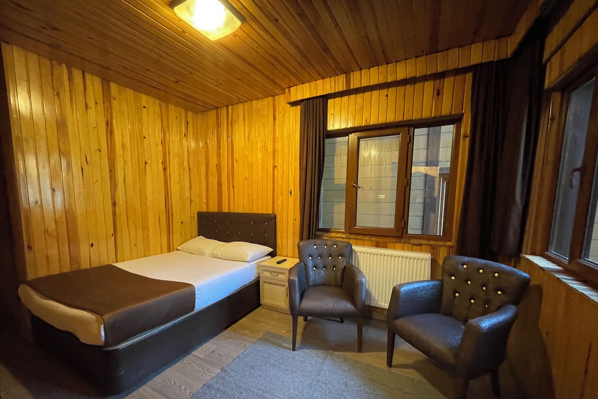 A wooden-paneled room with a bed, two armchairs, a nightstand, and a window with curtains.