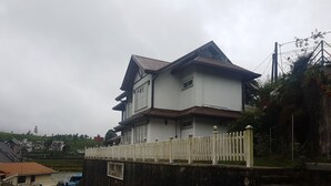 A two-story house with a white exterior and a brown roof, surrounded by a white picket fence and a stone wall. - Villa lake view (Nuwara Eliya)