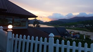 A view of a riverside town with a white picket fence in the foreground, a building with a dark roof, and mountains in the background. - Villa lake view (Nuwara Eliya)