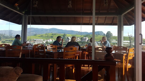 A restaurant with wooden furniture and large windows offering a view of green hills and buildings. - Villa lake view (Nuwara Eliya)