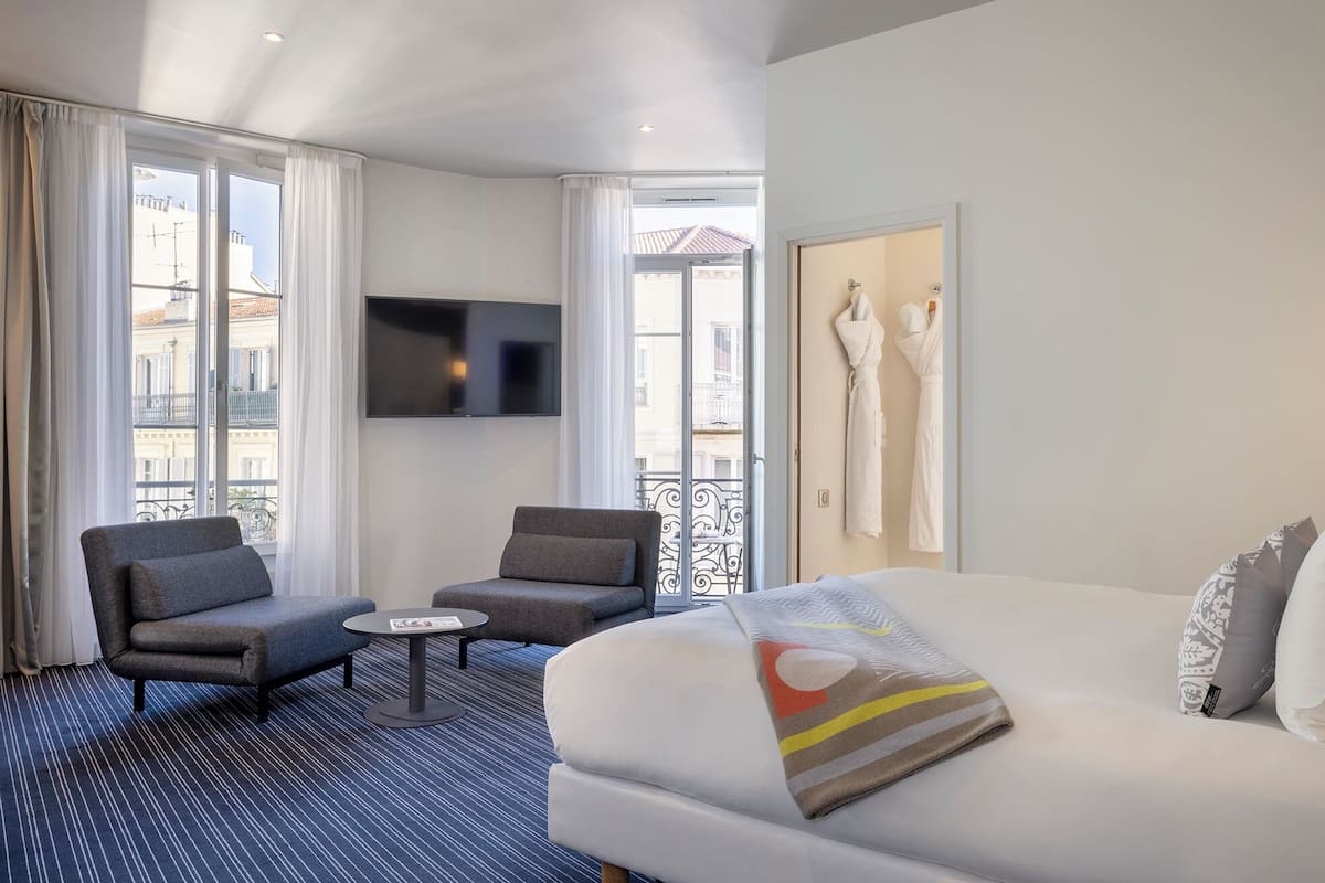 A hotel room with a large bed, two armchairs, a small table, a TV, and a balcony with a view of buildings.