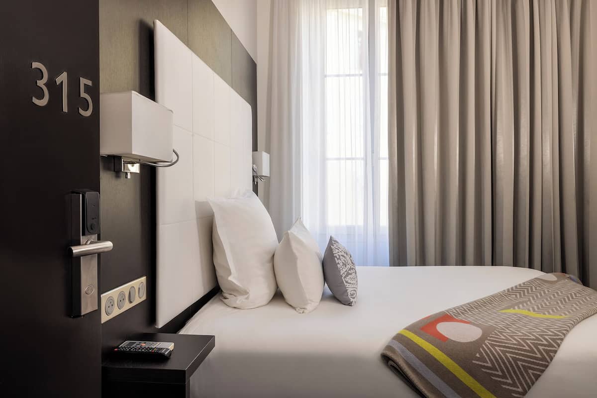 A modern hotel room with a large bed, a headboard, a bedside table with a remote control, and a number "315" on the wall.