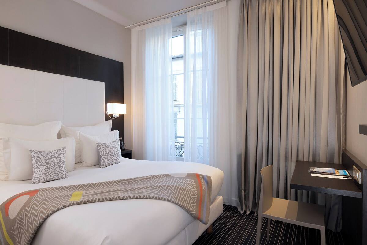 A hotel room with a large bed, a desk, and a view of the city through sheer curtains.