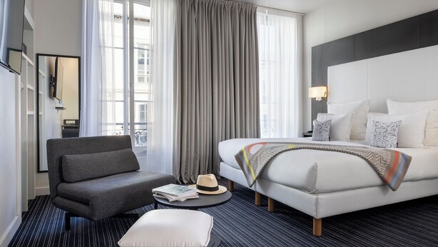 A modern hotel room with a bed, a grey armchair, a small round table with a hat and a magazine, and a large window with curtains. - Hôtel 66 (Nice)