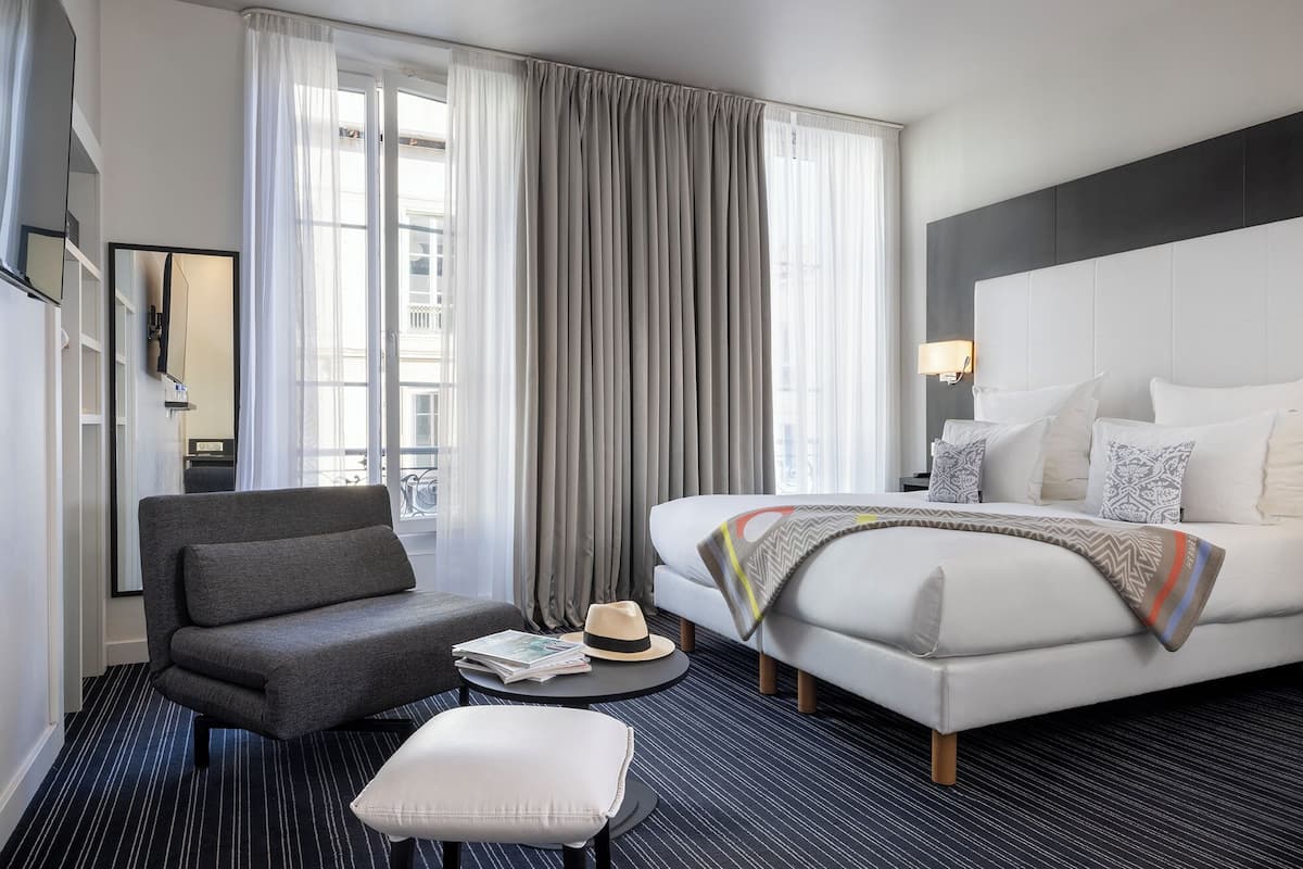 A modern hotel room with a bed, a grey armchair, a small round table with a hat and a magazine, and a large window with curtains.