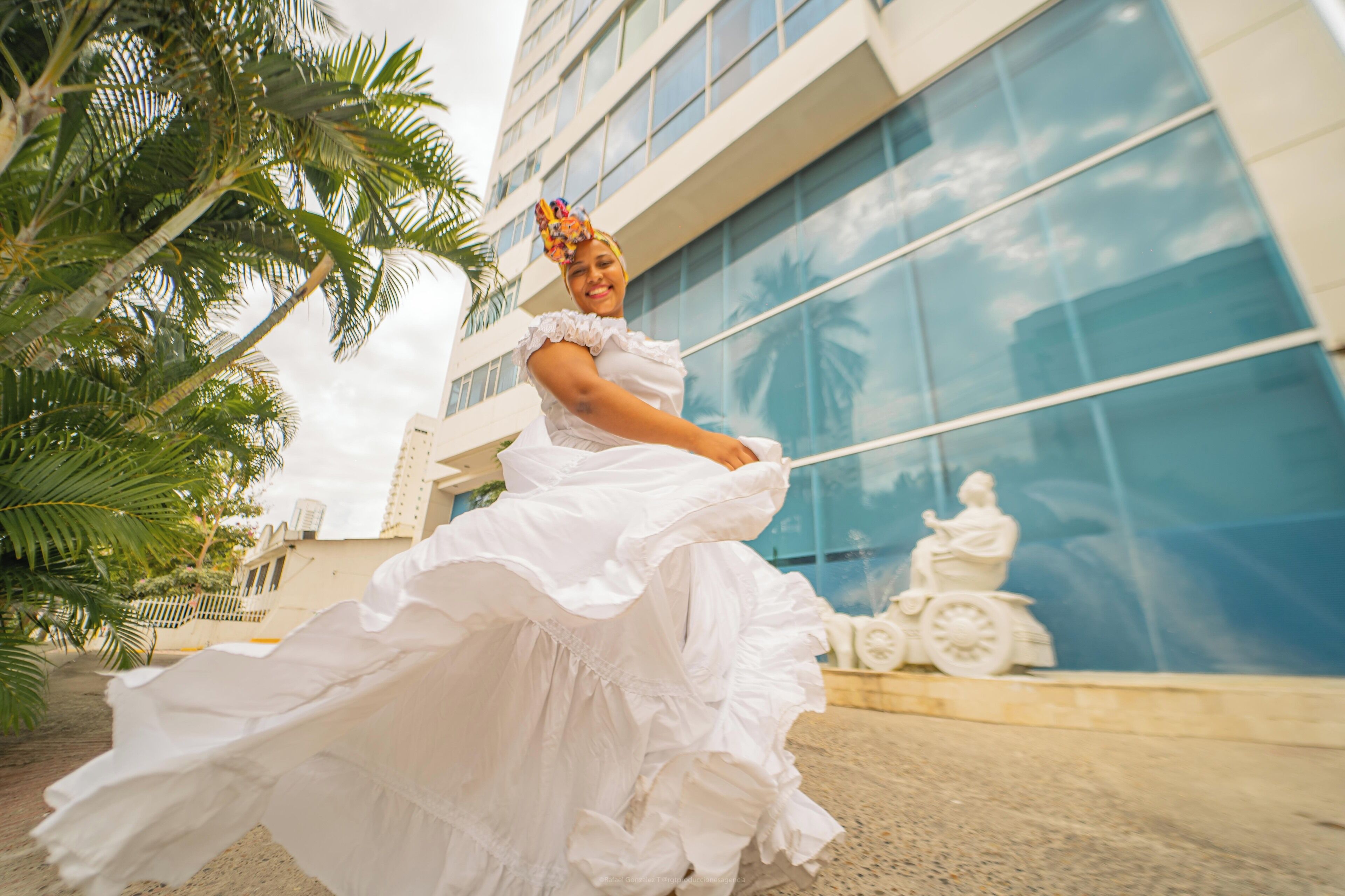 A person in a white dress with ruffled sleeves, standing in front of a modern building with glass windows and a statue.