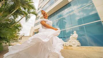 A person in a white dress with ruffled sleeves, standing in front of a modern building with glass windows and a statue.
