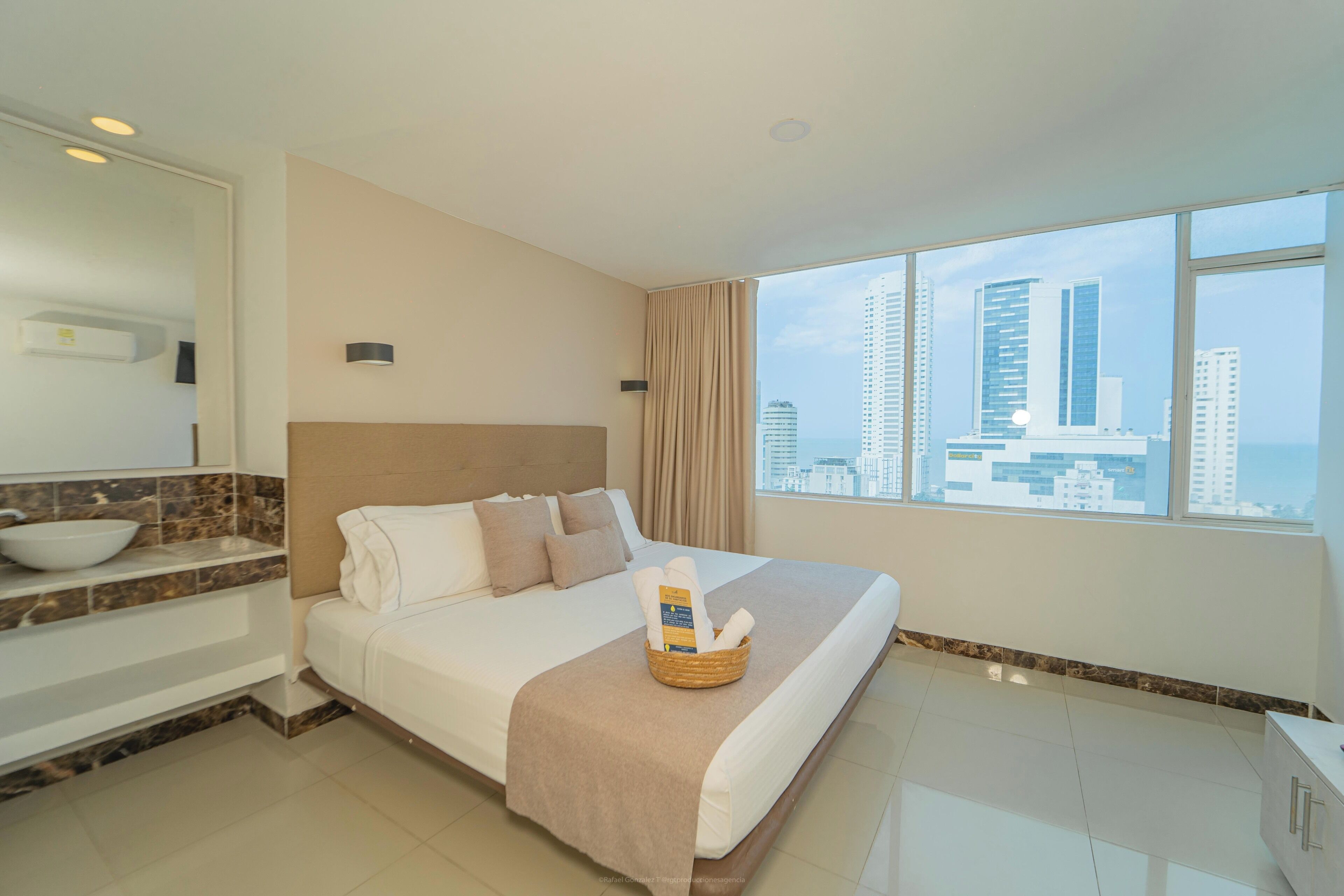 A hotel room with a bed, a bathroom with a sink, and a large window with a city view.