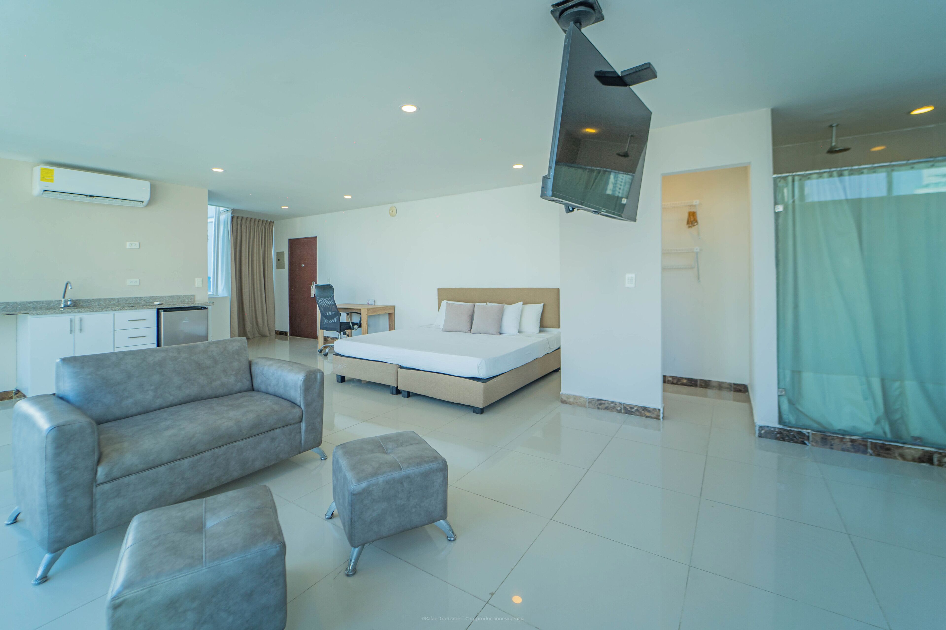 A modern hotel room with a bed, a sofa, an ottoman, a kitchenette, and a shower area.