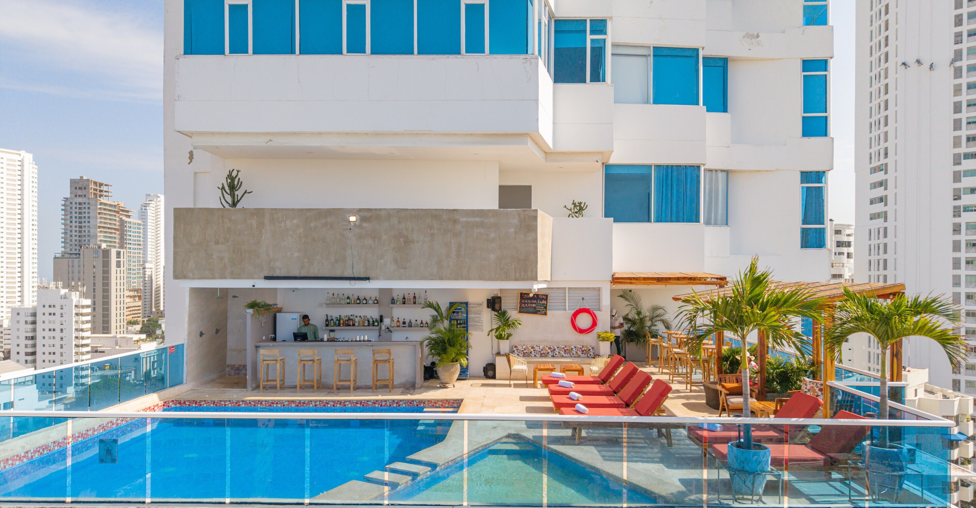 A rooftop pool area with lounge chairs, a bar, and a view of surrounding high-rise buildings.