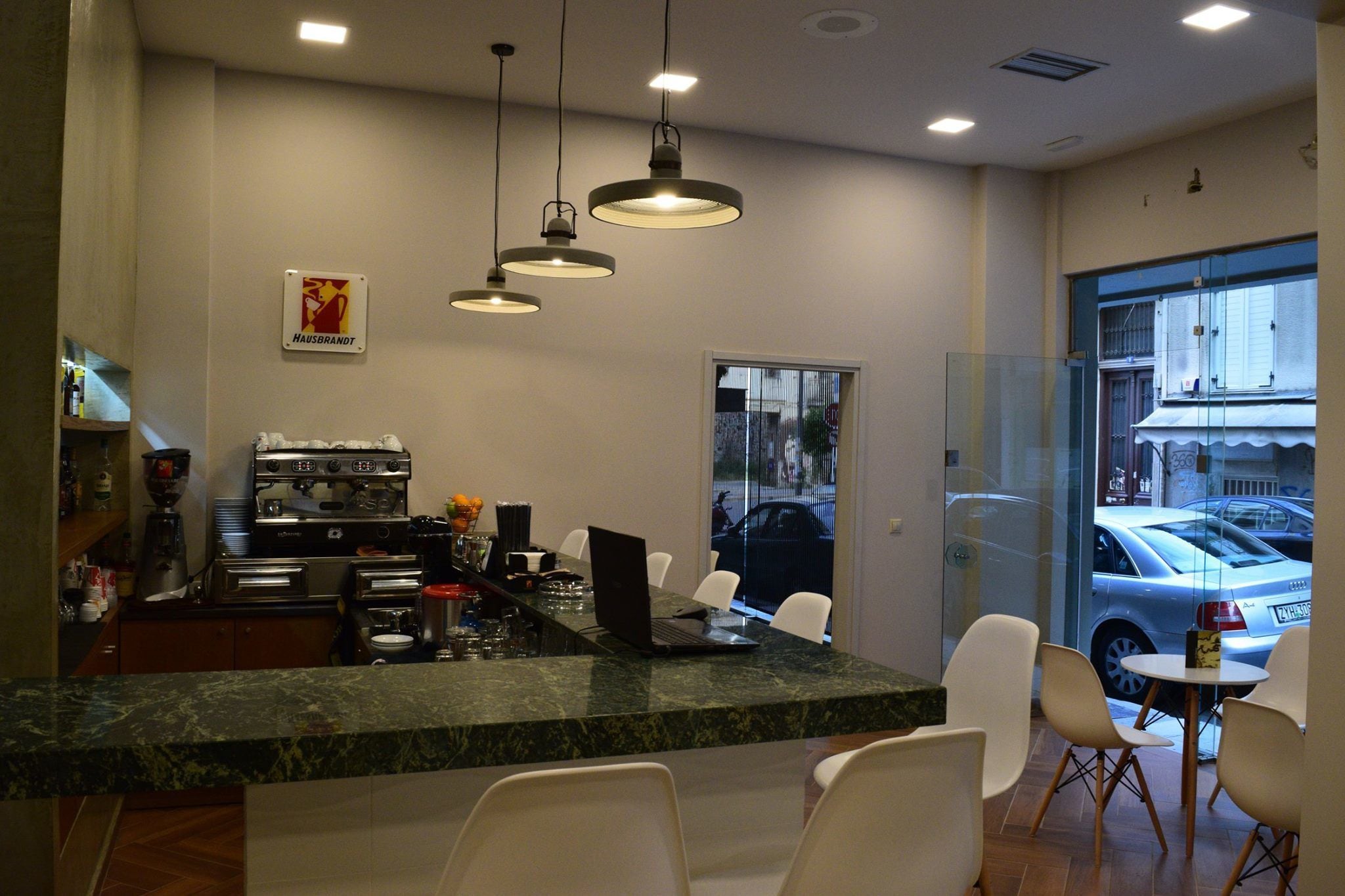 Eine moderne Küche mit Marmorarbeitsplatte, einer Kaffeemaschine und einem Laptop auf der Arbeitsfläche. Weiße Stühle umgeben einen kleinen Tisch und eine Glastür bietet Blick auf die Straße.