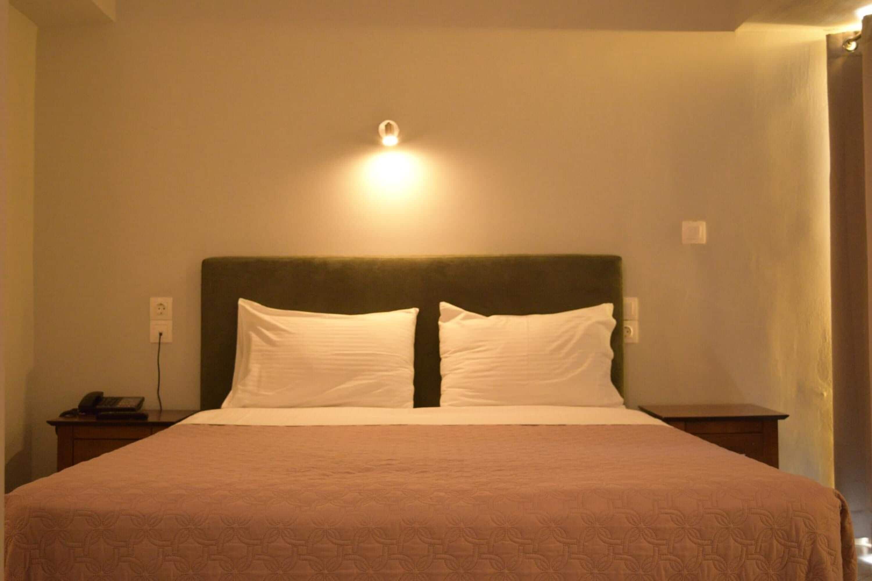 Una cama bien hecha con cabecera, una mesita de noche con teléfono, y una lámpara fijada a la pared.