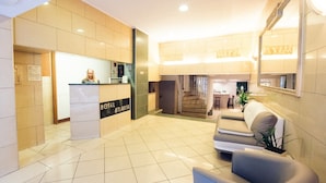 A hotel reception area with a reception desk, a staircase leading to an upper floor, and a seating area with a plant.