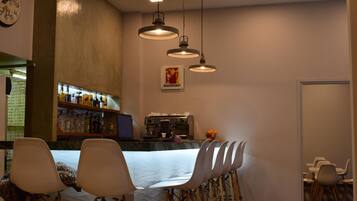 A bar area with a counter, shelves with bottles, and a coffee machine. There are chairs and a table in the foreground.