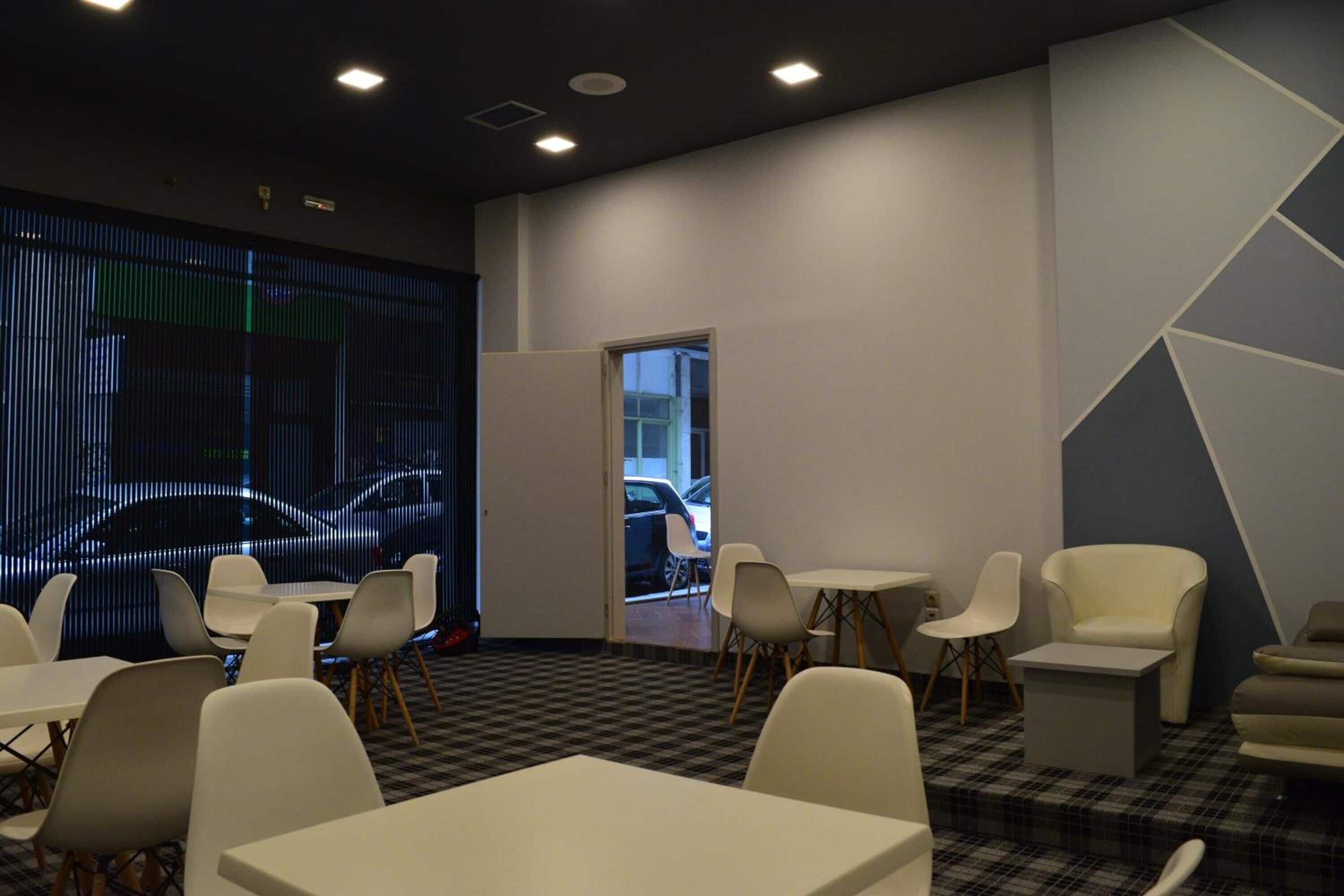 A modern waiting area with a checkered floor, white tables, and chairs, a large digital screen displaying a car, and a door leading outside.