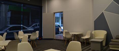 A modern waiting area with a checkered floor, white tables, and chairs, a large digital screen displaying a car, and a door leading outside.