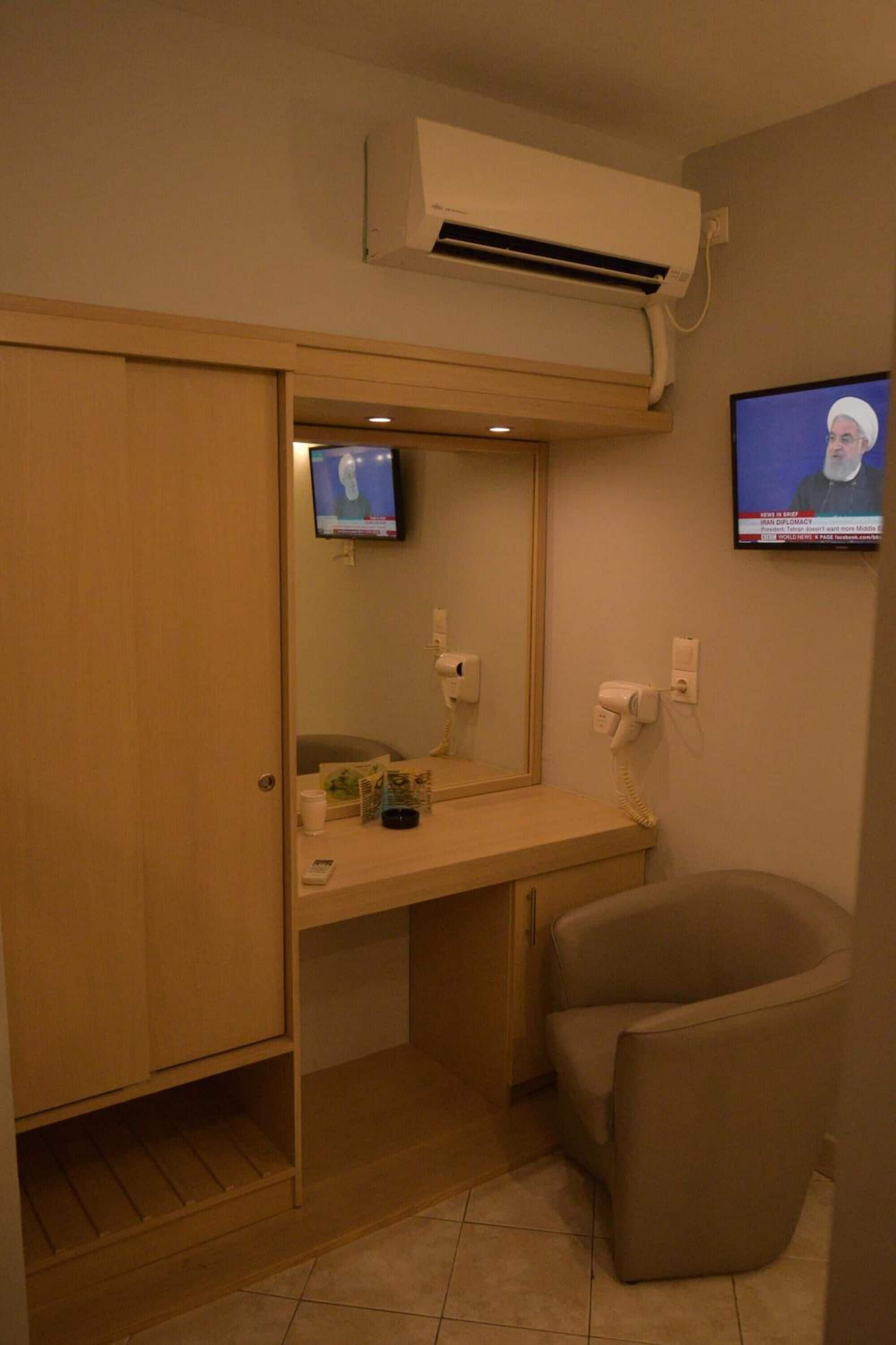 A hotel room with a wooden wardrobe, a small desk with a mirror, a television mounted on the wall, and a beige armchair.