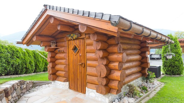 A log cabin with a wooden door, stone foundation, and a small garden area with a stone wall and greenery. - Hotel Kristall (Hermagor-Pressegger See)