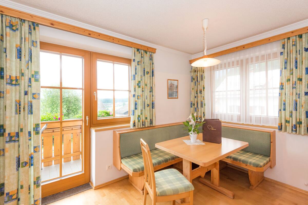 A dining area with a wooden table and bench seating, large windows with curtains, and a view of greenery outside.