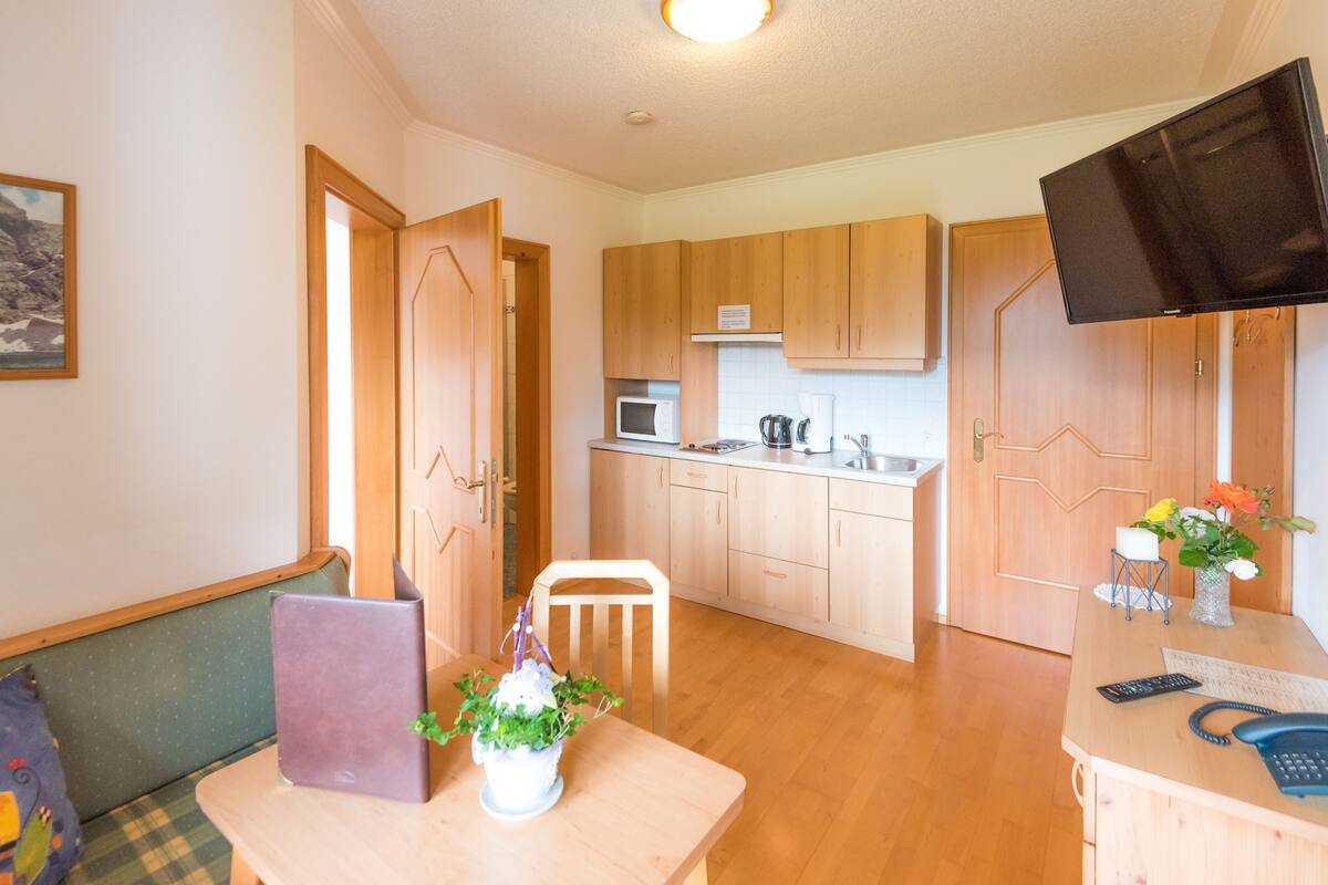 A compact kitchen with wooden cabinets, a microwave, and a coffee maker. There's a small table with a laptop and a plant, and a TV mounted on the wall.