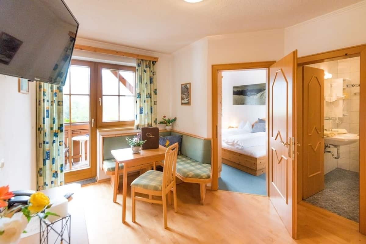 A compact room with a bed, a small table with chairs, a window with curtains, and a bathroom with a sink and mirror.