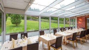 A restaurant with tables set for dining, large windows offering a view of a green lawn and trees. - Hotel Kristall (Hermagor-Pressegger See)