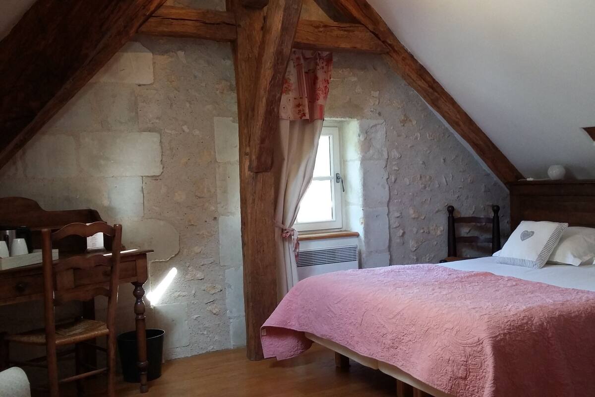 A bedroom with a sloped ceiling, a wooden bed with white pillows, a pink blanket, and a rustic stone wall.