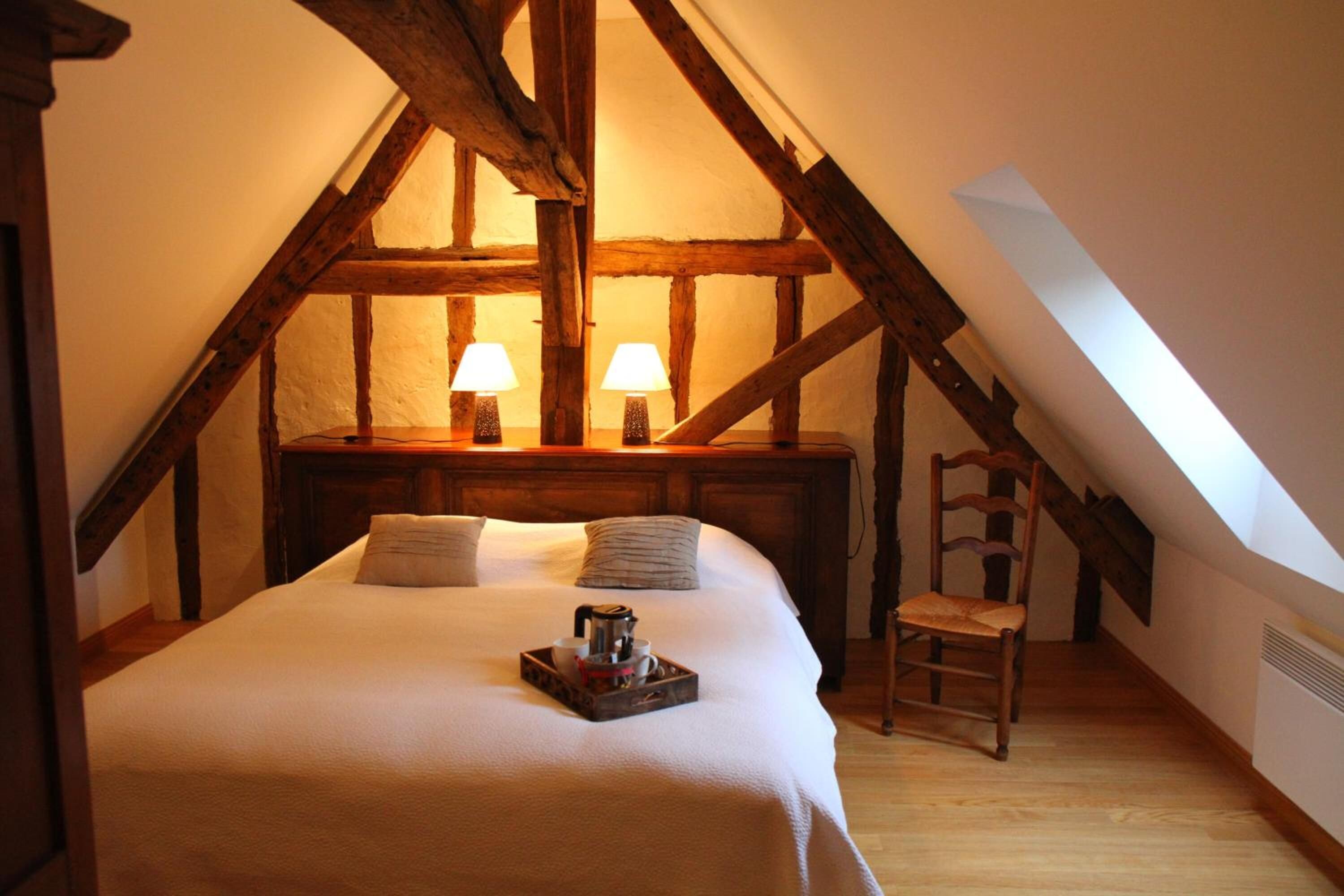 A cozy attic bedroom with a high ceiling, exposed wooden beams, a bed with pillows, two bedside lamps, and a small table with a tray.