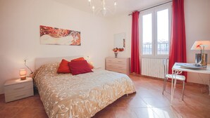 Une chambre à coucher comprenant un lit, une table de chevet, un bureau, une chaise, une fenêtre avec des rideaux rouges et un lustre.