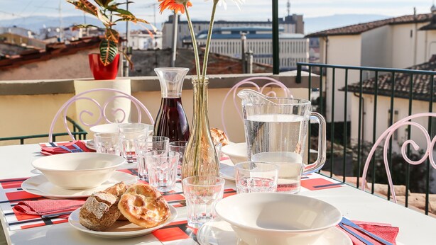 Une table dressée avec des assiettes, des verres, une carafe et un vase rempli de fleurs, donnant sur un paysage urbain.