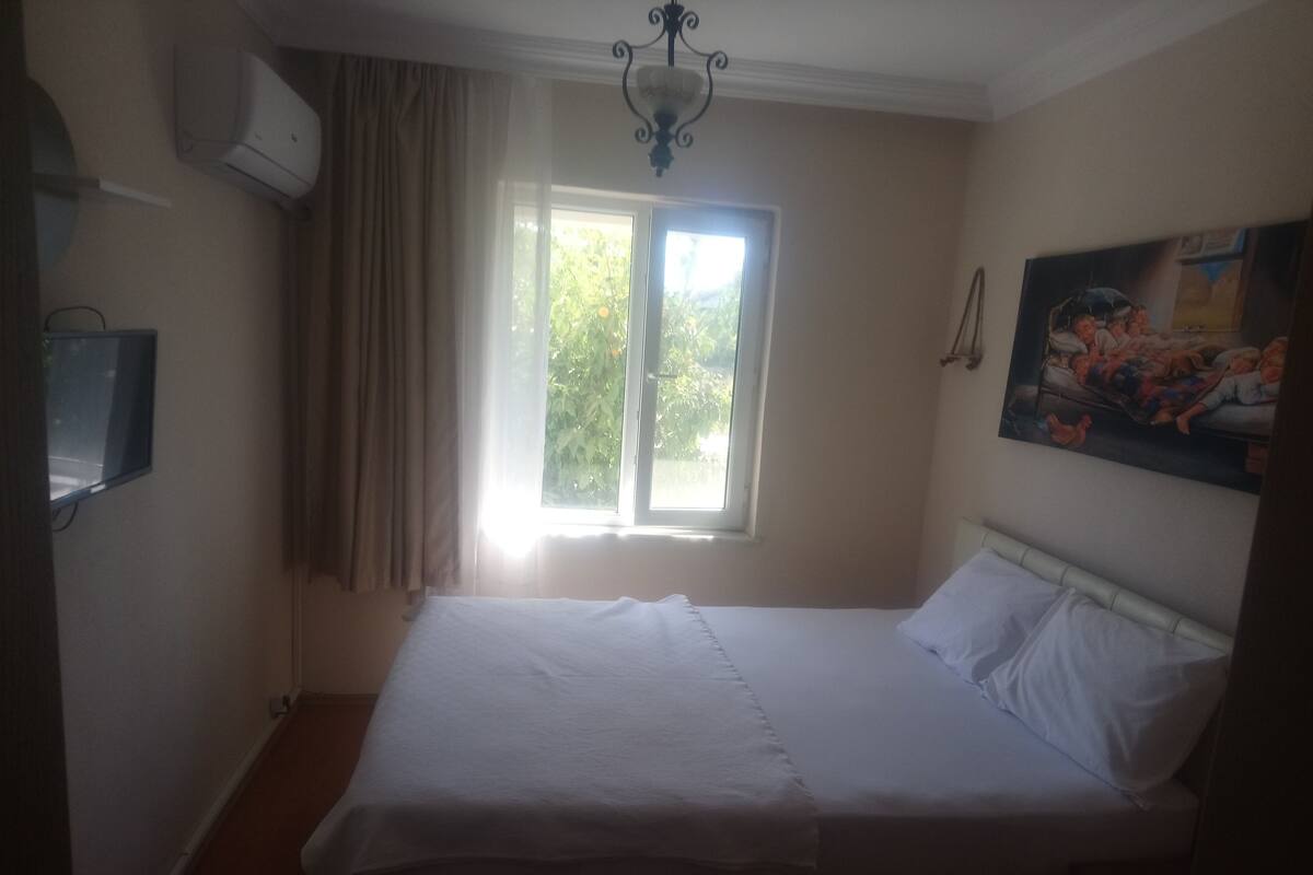A bedroom with a bed, a window with curtains, a painting on the wall, and an air conditioning unit.