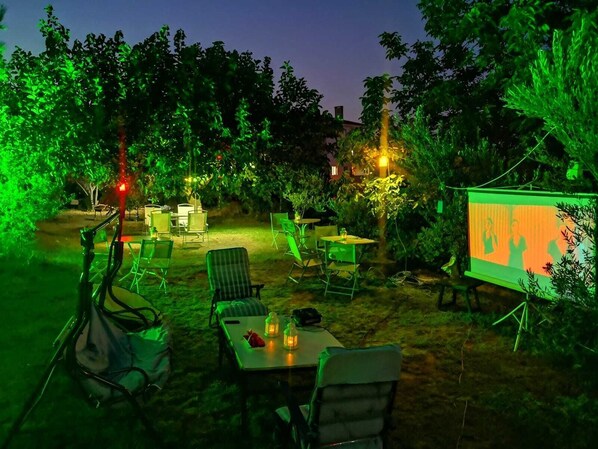 An outdoor seating area with tables and chairs, surrounded by trees and a projection screen displaying an image. - Bahceli Konak (Eceabat)