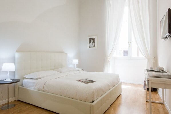 Un dormitorio ordenado con cama, mesitas de noche, lámpara, un cuadro enmarcado y una ventana con cortinas.
