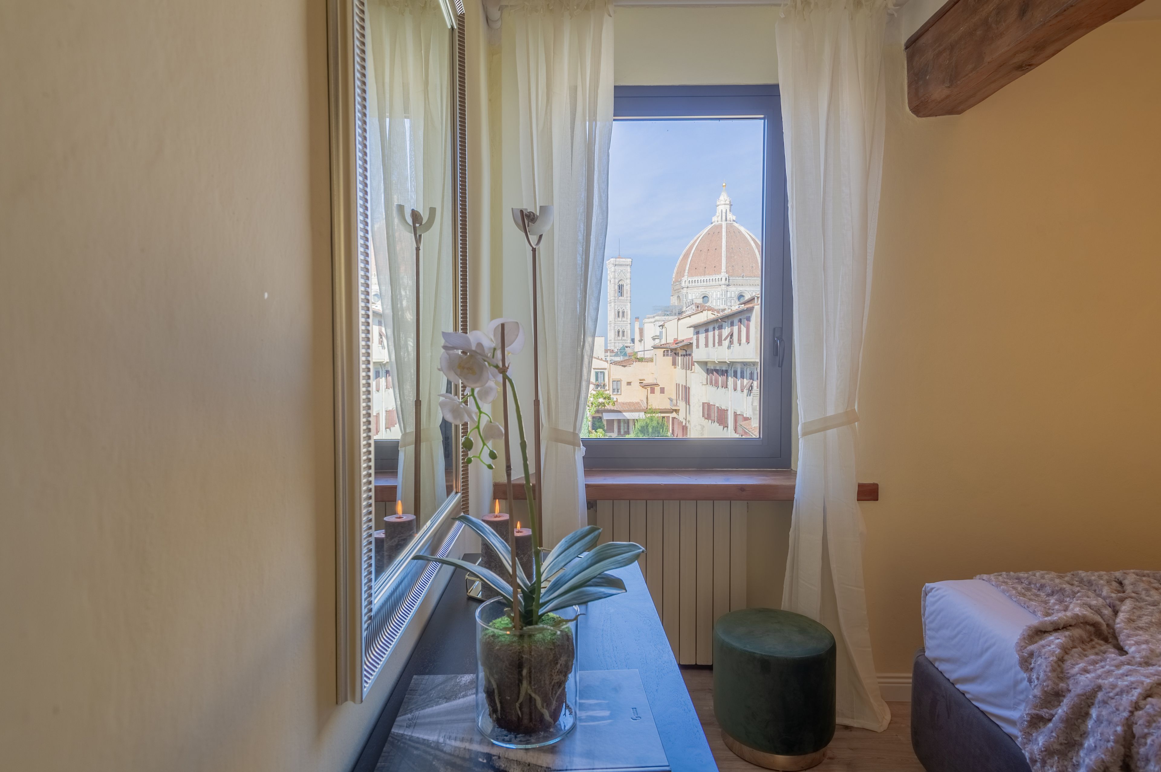 A room with a window overlooking a cityscape, a bed, a small table with a plant, and a mirror.